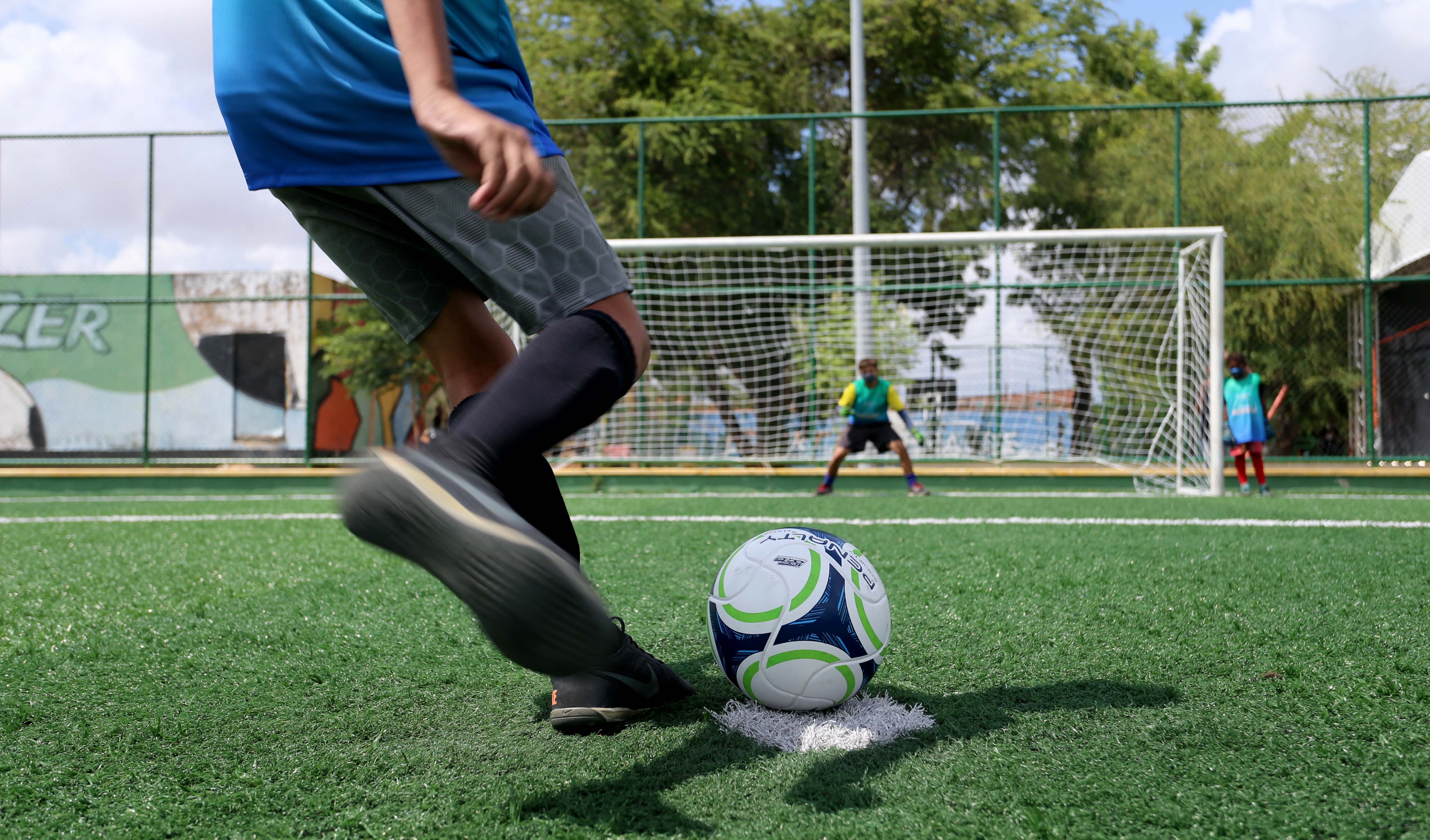 a foto mostra uma criança de costas chutando uma bola e, ao fundo, outra criança está no gol esperando para defender